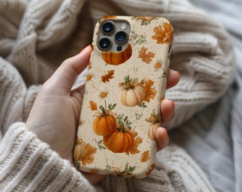 May include: A beige phone case featuring a fall-themed pattern. The design includes orange pumpkins, brown leaves, and white daisies. The case is held in a hand, showcasing the detailed print and the phone's camera lenses.