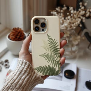 May include: A white phone case with a fern leaf design. The case has a camera lens cutout and a cream background.