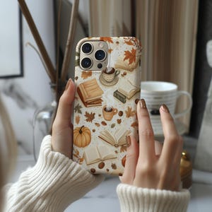 Puede incluir: Una funda de teléfono con un fondo crema, decorada con ilustraciones de libros, tazas de café, calabazas y hojas de otoño en marrón, naranja y verde. La funda la sostiene una persona que lleva un jersey color crema.
