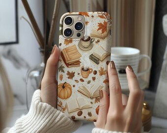 May include: A phone case featuring a fall-themed design. The case has a cream background with illustrations of books, coffee cups, pumpkins, and autumn leaves in shades of brown, orange, and green. The case is held by a person wearing a cream-colored sweater.