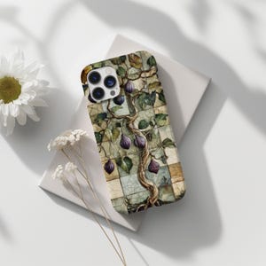 May include: A smartphone case featuring a fig tree design. The case has a mosaic tile background with a tree trunk, green leaves, and purple figs. The case is resting on a white surface with a daisy and small white flowers.