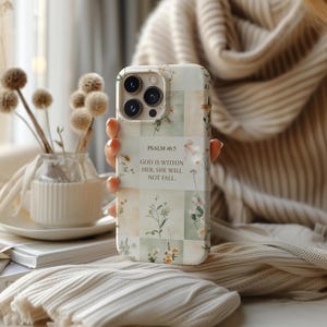 May include: A floral patterned phone case with the text "PSALM 46:5 GOD IS WITHIN HER, SHE WILL NOT FALL." The case features a collage of pastel green and cream squares with delicate flower illustrations. The phone case is held in a person's hand.