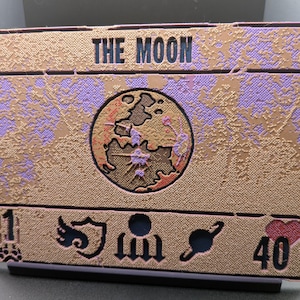 May include: A rectangular card with the text "THE MOON" at the top. The card features a textured gold and purple design with a central circular image of a globe. The bottom has symbols and the numbers 1 and 40. The card is on a black stand.