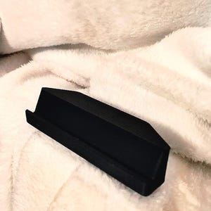 May include: A black, angled phone stand with a textured surface. The stand is designed to hold a phone upright and is set against a soft, white, textured background.