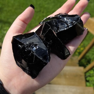 Peut inclure: Trois morceaux de pierre d'obsidienne noire brute avec une texture brillante semblable à du verre et des bords de fracture conchoïdaux tranchants. Les pierres sont opaques, d'un noir profond, avec des surfaces lisses et réfléchissantes.