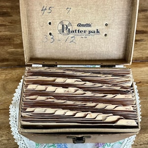 May include: A brown vintage record storage case with the text "Amlite Platter-pak Phonograph Record Case No. 700" and the numbers "45 7 33-1/2" written on the lid. The case is open and has 50 slots for records.