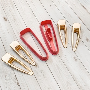 May include: Four gold hair clips and two pink plastic hair clip cutters on a light wood surface.