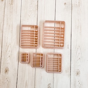 May include: Set of five pink plastic cookie cutters shaped like a grid. The cutters are different sizes and are arranged in a row on a light wood surface.