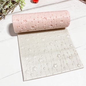 May include: A pink rolling pin with a star and vine pattern is shown rolled out on a white fondant sheet with the same pattern. The rolling pin is used to create decorative patterns on fondant for cakes and other desserts.