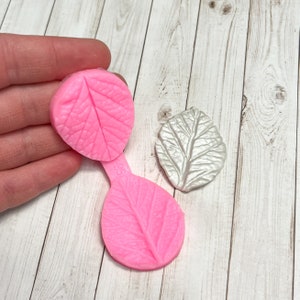 May include: Two pink silicone molds for making leaf shapes. The molds are connected by a thin strip of silicone. The molds are on a white wooden surface.
