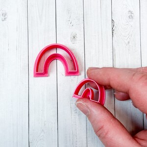 May include: Two pink plastic rainbow-shaped cookie cutters. The larger cutter is on a white wooden surface. The smaller cutter is being held by a hand.