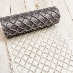 May include: A gray rolling pin with a geometric pattern embossed on the surface. The rolling pin is resting on a white surface with a similar pattern embossed on it.