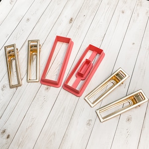 May include: Four gold hair clips and two pink plastic hair clip molds. The molds are rectangular with a cut-out section in the middle. The hair clips are all the same size and shape, with a rectangular design.
