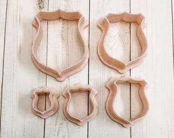 May include: Set of five plastic cookie cutters in the shape of a shield. The cutters are different sizes and are arranged in a row on a light wood surface.