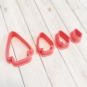 May include: Four pink plastic cookie cutters shaped like Christmas trees, arranged in descending size order from left to right.