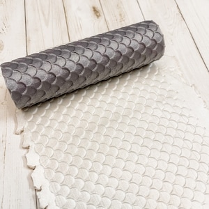 May include: A gray silicone rolling pin with a scale pattern, laying on a white silicone baking mat with a scale pattern.