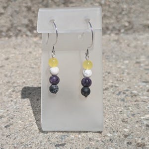 May include: A pair of dangle earrings featuring silver-toned hooks and a vertical arrangement of four beads each. The beads consist of a translucent yellow sphere, a white sphere, a purple sphere, and a black sphere with a white crackle pattern.