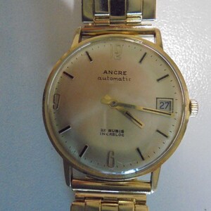 May include: A gold-toned wristwatch with a round face and a matching gold band. The watch face displays the text "ANCRE automatic" and "25 RUBIS INCABLOC". The date is visible at the 3 o'clock position.