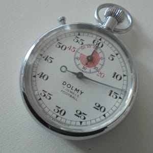 Puede incluir: Un cronómetro plateado con esfera blanca, números negros y esfera interior roja. El reloj indica "DOLMY 7 JEWELS FOOTBALL". Las manecillas señalan aproximadamente 40 segundos. El reloj tiene una corona y un botón superior.