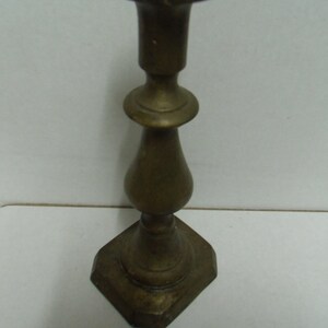 Puede incluir: Un candelero de latón vintage con base hexagonal y parte superior acampanada. El candelero tiene un diseño clásico con una sección media bulbosa y un tallo delgado. El latón tiene una pátina ligeramente envejecida.