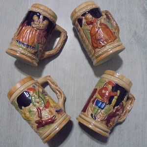 Può includere: Quattro boccali da birra in ceramica con scene in rilievo dettagliate e manici ornati. I boccali presentano immagini di persone in abiti tradizionali, con una combinazione di colori beige e marrone. I boccali sono disposti su una superficie grigio chiaro.