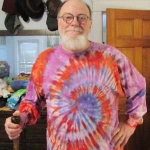 May include: A man with a white beard is wearing a long-sleeved tie-dye shirt with a red, blue, and purple swirl pattern. He is holding a wooden walking stick.