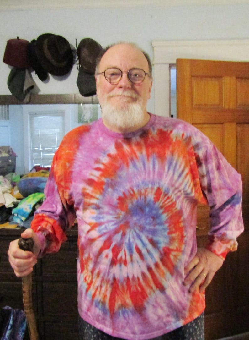 May include: A man with a white beard is wearing a long-sleeved tie-dye shirt with a red, blue, and purple swirl pattern. He is holding a wooden walking stick.