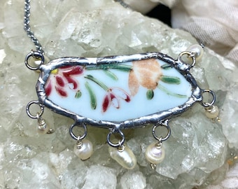 May include: A delicate necklace featuring a vintage porcelain pendant with a floral design in shades of pink, orange, and green. The pendant is surrounded by a silver frame and hangs from a silver chain with pearl accents.