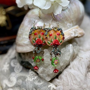 May include: A pair of dangle earrings with a round, painted glass charm featuring red roses and green leaves. The charms are attached to a gold-toned metal chain with green and red glass beads.
