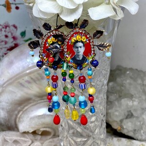 May include: A pair of colorful dangle earrings featuring a portrait of Frida Kahlo in a red oval frame. The earrings are made with a variety of beads, including red, blue, green, yellow, and white.
