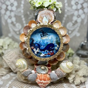 May include: A handcrafted nautical-themed decorative piece featuring a central circular diorama with a whale and ship scene. The diorama is surrounded by various seashells in shades of tan, orange, and white. The piece is topped with white flowers.
