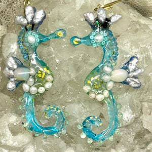 May include: A pair of blue seahorse earrings with white pearl accents and gold hooks. The seahorses are decorated with gold glitter and white pearls.