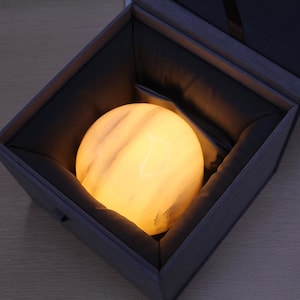 May include: A spherical, glowing lamp with a smooth, marbled surface featuring warm yellow and white tones. The orb is nestled inside a dark gray, square-shaped presentation box with a fabric-lined interior, creating a soft, diffused light.