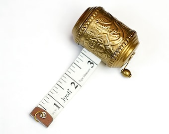May include: A vintage-style brass tape measure with a floral design. The tape measure extends to 3 inches, with the word "Jyoti" printed on the white tape. The brass casing has an ornate, embossed pattern.