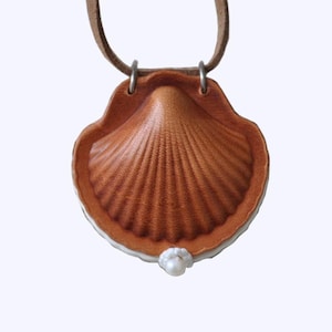 May include: A brown leather pendant necklace in the shape of a scallop shell. The shell features detailed ridged texture and a white border. A small pearl is attached at the bottom. The pendant hangs from a brown leather cord.