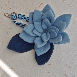 Op de afbeelding: Een spijkerstof bloem sleutelhanger met een lichtblauwe bloem en donkerblauwe bladeren. De bloem heeft een kleine, donkere kraal in het midden. De sleutelhangerband is gevlochten met witte, blauwe en marineblauwe draden.
