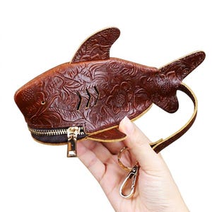 Può includere: Un portamonete in pelle marrone scuro a forma di squalo, caratterizzato da un intricato motivo floreale in rilievo su tutta la superficie. La pochette include una chiusura a zip in metallo dorato, tre piccole fessure e un cinturino da polso.
