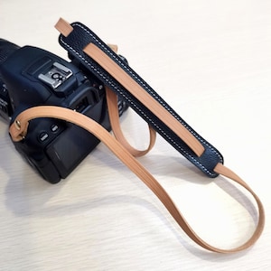 Peut inclure: Un sangle de caméra en cuir noir avec un accent en cuir beige. La sangle est attachée à un appareil photo noir.