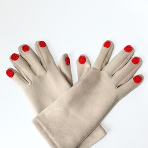 Könnte beinhalten: Ein Paar beigefarbene Handschuhe mit kleinen, runden roten Perlenverzierungen an den Fingerspitzen, die das Aussehen manikürter Fingernägel nachahmen. Die Handschuhe bestehen aus einem glatten, einfarbigen Stoffmaterial.
