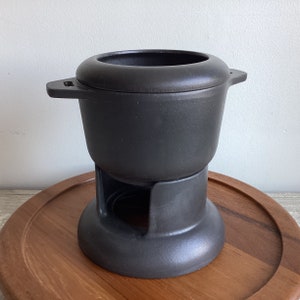 May include: Black cast iron fondue pot with a stand. The pot has two handles and a round base. The stand has a circular opening in the centre.