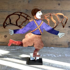 May include: A whimsical metal sculpture of a person with wings, wearing a purple sweater, brown trousers, and red boots. The sculpture is painted with details and has a rustic, handmade look.