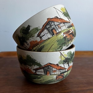 Peut inclure: Deux bols en céramique avec un motif peint à la main représentant une maison dans un paysage de campagne. Les bols sont empilés l'un sur l'autre. Le motif représente une maison blanche avec un toit brun, des arbres verts et une clôture brune.