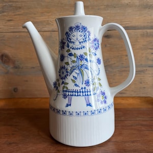 以下が含まれることがあります： 白のセラミックコーヒーポット。青と紫の花柄のデザイン。花冠と花束を持つ様式化された人物が描かれています。ポットは長い注ぎ口、ハンドル、蓋が付いています。