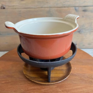 Puede incluir: Una pequeña olla de fondue naranja con interior blanco y dos asas. La olla se encuentra sobre un soporte de metal negro con base de madera. La olla está diseñada para cocinar y servir fondue.