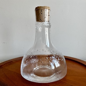 May include: A clear glass decanter with a textured surface and a cork stopper. The decanter has a wide base and a narrow neck.