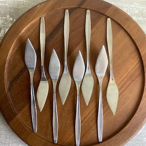 Pode incluir: Sete facas de manteiga prateadas com pontas pontiagudas são dispostas em uma superfície de madeira. As facas são todas do mesmo tamanho e forma, e são dispostas em uma fileira.