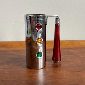 Könnte beinhalten: Ein verchromter Cocktailshaker mit rotem Griff und drei farbigen Lichtern: rot, gelb und grün. Die Worte "Danger", "Caution" und "Safe" sind darunter eingraviert. Der Shaker steht auf einer Holzoberfläche.