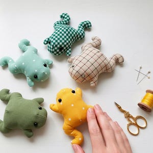 Peut inclure: Plusieurs peluches en forme de grenouille faites &agrave; la main, de diff&eacute;rentes couleurs et motifs, dont du vichy vert, du bleu clair &agrave; pois blancs et du jaune. Des fournitures de couture, dont des ciseaux, des &eacute;pingles et du fil, sont &eacute;galement visibles.