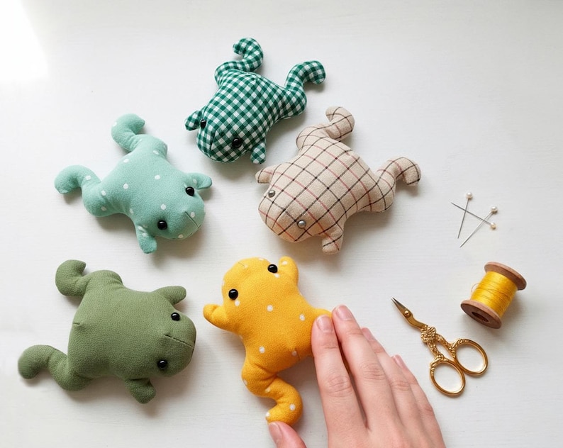 Peut inclure: Plusieurs peluches en forme de grenouille faites &agrave; la main, de diff&eacute;rentes couleurs et motifs, dont du vichy vert, du bleu clair &agrave; pois blancs et du jaune. Des fournitures de couture, dont des ciseaux, des &eacute;pingles et du fil, sont &eacute;galement visibles.