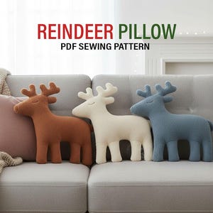 May include: Three reindeer-shaped cushions in brown, white, and blue are arranged on a grey sofa. A pink cushion and a beige throw are also present. The text "REINDEER PILLOW" and "PDF SEWING PATTERN" are displayed above the cushions.